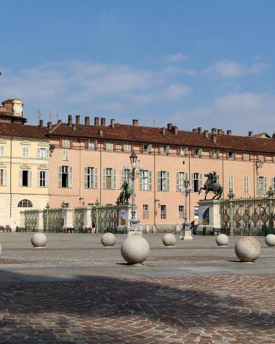 The image shows a large, paved public square with distinctive spherical stone bollards in the foreground, flanked by historic buildings including a prominent church with a dome on the left and a long pinkish palace behind an ornate iron gate featu