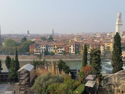 A panoramic view of Verona, Italy, showing the Adige River, Ponte Pietra bridge, city skyline with prominent towers (including Torre dei Lamberti and Campanile of San Zeno), and in the foreground, ancient Roman ruins on the hillside — specifically