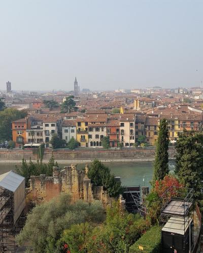 A panoramic view of Verona, Italy, showing the Adige River, Ponte Pietra bridge, city skyline with prominent towers (including Torre dei Lamberti and Campanile of San Zeno), and in the foreground, ancient Roman ruins on the hillside — specifically