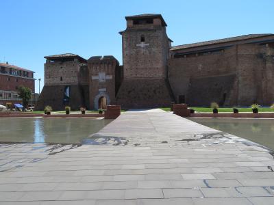 A low-angle view of a large, 15th-century brick fortress with multiple towers and a central arched entrance, situated behind a modern paved plaza and a shallow reflecting pool under a clear blue sky.
