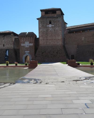 A low-angle view of a large, 15th-century brick fortress with multiple towers and a central arched entrance, situated behind a modern paved plaza and a shallow reflecting pool under a clear blue sky.