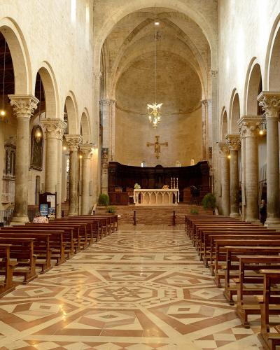 The interior of a Romanesque-style cathedral featuring a geometrically patterned marble floor, rows of wooden pews, stone columns with carved capitals, and an altar area at the far end under a large arch.