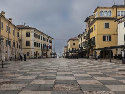 A wide, paved town square with a distinctive black-and-white checkered stone pattern, flanked by yellow and cream-colored buildings with shuttered windows, closed umbrellas, and flags on poles under an overcast sky.