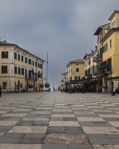 A wide, paved town square with a distinctive black-and-white checkered stone pattern, flanked by yellow and cream-colored buildings with shuttered windows, closed umbrellas, and flags on poles under an overcast sky.
