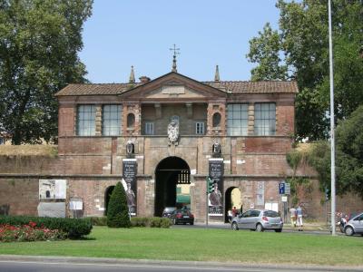 The image shows a large, brick-and-stone city gate with a central archway, flanked by smaller arches, featuring banners that read "Piazza Lucca" and a pediment inscribed with "LIBERTAS," set against a grassy foreground and trees.