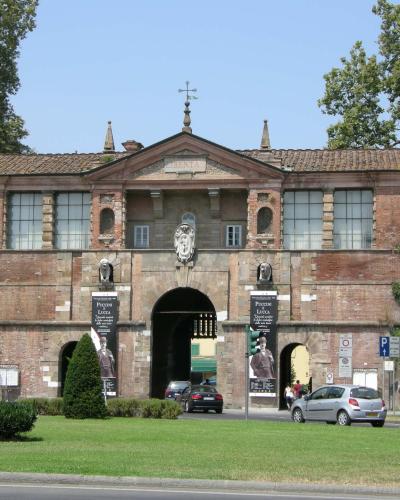 The image shows a large, brick-and-stone city gate with a central archway, flanked by smaller arches, featuring banners that read "Piazza Lucca" and a pediment inscribed with "LIBERTAS," set against a grassy foreground and trees.