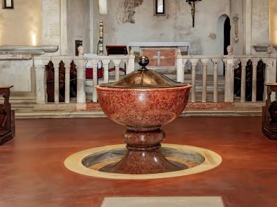 The image shows the interior of a church featuring a large, ornate baptismal font made of reddish veined marble with a metal lid, set on a terracotta-colored floor with a circular stone surround, and a white stone altar railing in the background.