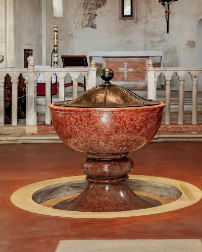 The image shows the interior of a church featuring a large, ornate baptismal font made of reddish veined marble with a metal lid, set on a terracotta-colored floor with a circular stone surround, and a white stone altar railing in the background.