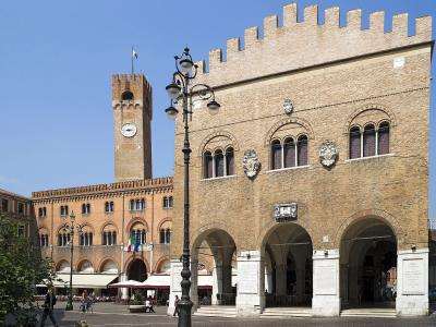 The image shows a sunlit Italian piazza featuring the Palazzo dei Trecento with its distinctive crenellated facade and arched loggia on the right, and the Torre Civica (clock tower) of the Palazzo della Ragione in the background, with outdoor cafe