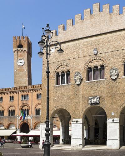 The image shows a sunlit Italian piazza featuring the Palazzo dei Trecento with its distinctive crenellated facade and arched loggia on the right, and the Torre Civica (clock tower) of the Palazzo della Ragione in the background, with outdoor cafe
