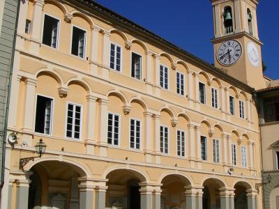 A large, pale yellow, multi-story building with a long arcade of arches on the ground floor and a clock tower topped with a cross, situated in a paved public square under a clear blue sky.
