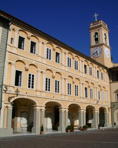 A large, pale yellow, multi-story building with a long arcade of arches on the ground floor and a clock tower topped with a cross, situated in a paved public square under a clear blue sky.