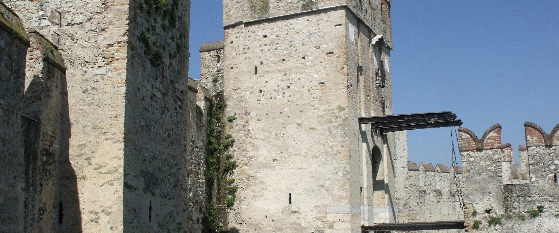 A stone medieval fortress with crenellated towers, a wooden drawbridge over a moat, and a small boat passing through the water gate under a clear blue sky.
