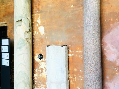 The image shows an interior museum or exhibition space with three ancient stone artifacts (two columns and a rectangular slab) displayed against a weathered, ochre-colored wall, with modern informational signage visible on the left.