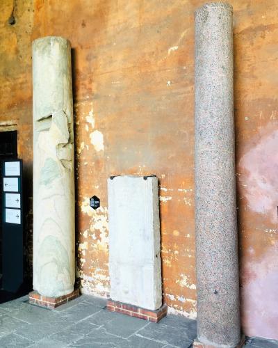 The image shows an interior museum or exhibition space with three ancient stone artifacts (two columns and a rectangular slab) displayed against a weathered, ochre-colored wall, with modern informational signage visible on the left.