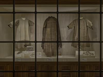 A display case viewed through a grid-patterned glass window, containing three historical religious garments (tunics and a mantle) and associated artifacts with Italian labels identifying them as belonging to St. Francis and St. Clare.