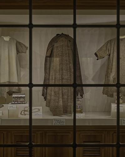 A display case viewed through a grid-patterned glass window, containing three historical religious garments (tunics and a mantle) and associated artifacts with Italian labels identifying them as belonging to St. Francis and St. Clare.