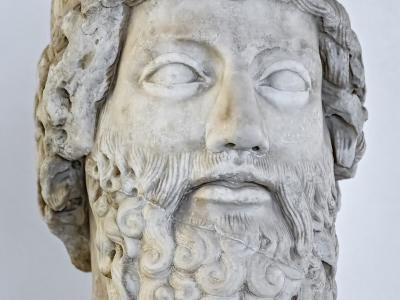 A marble bust of a bearded male figure with curly hair and beard, displayed on a white pedestal against a plain white background.