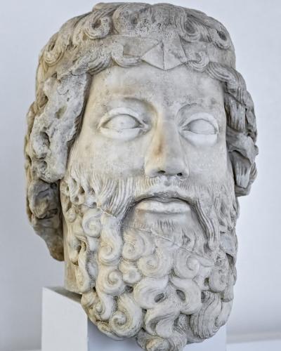 A marble bust of a bearded male figure with curly hair and beard, displayed on a white pedestal against a plain white background.