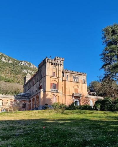 A large, terracotta-colored castle-like building with crenellated towers and Gothic-Revival architecture, situated on a grassy slope with a rocky hillside in the background under a clear blue sky.
