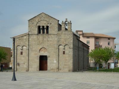 A stone Romanesque church with a central gable, arched windows, a small bell tower, and a brown sign on the right reading "BASILICA SAN SIMPLICIO" in a paved plaza.