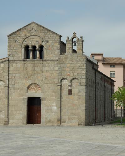 A stone Romanesque church with a central gable, arched windows, a small bell tower, and a brown sign on the right reading "BASILICA SAN SIMPLICIO" in a paved plaza.