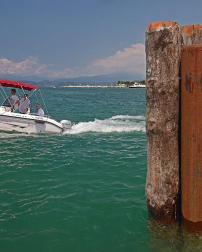 A white motorboat with a red canopy, labeled "PESCHIERA," moves across turquoise water past two wooden mooring posts in the foreground, with a distant shoreline and mountains under a partly cloudy sky.