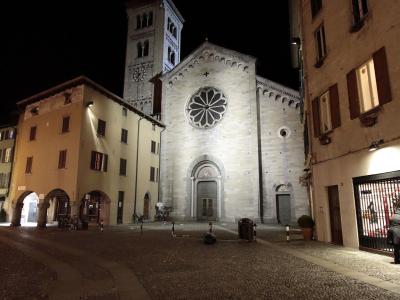 A night photograph of a large, light-colored stone Romanesque church with a prominent rose window and an adjacent tall bell tower, situated in a cobblestone piazza flanked by other buildings.