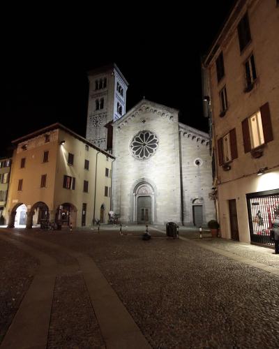 A night photograph of a large, light-colored stone Romanesque church with a prominent rose window and an adjacent tall bell tower, situated in a cobblestone piazza flanked by other buildings.