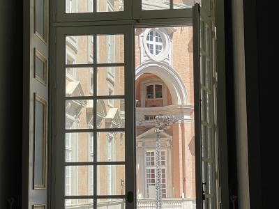A view from inside a room looking out through an open, multi-paned window onto the brick facade and courtyard of a large, ornate palace with classical architectural details.
