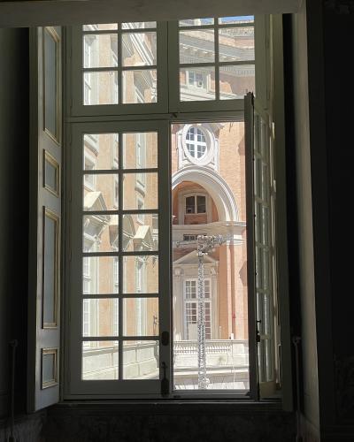 A view from inside a room looking out through an open, multi-paned window onto the brick facade and courtyard of a large, ornate palace with classical architectural details.