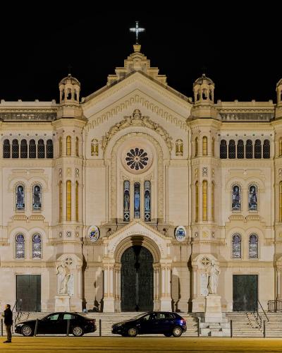 A brightly illuminated neoclassical cathedral facade at night, featuring a central rose window, flanking towers with cupolas, statues on pedestals, and cars parked along the street in front.