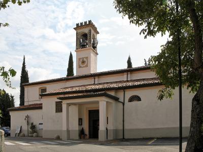 A white-walled church with a terracotta-tiled roof, a prominent square bell tower featuring a clock and open belfry, and a small portico entrance, set against a partly cloudy sky with surrounding trees.