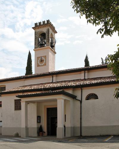 A white-walled church with a terracotta-tiled roof, a prominent square bell tower featuring a clock and open belfry, and a small portico entrance, set against a partly cloudy sky with surrounding trees.