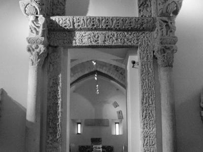 A black-and-white photograph of an ornate, carved stone portal with lion-head keystones and intricate relief work, displayed indoors within a museum or exhibition space.