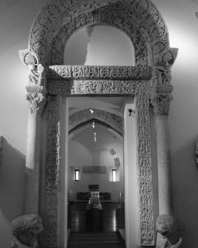 A black-and-white photograph of an ornate, carved stone portal with lion-head keystones and intricate relief work, displayed indoors within a museum or exhibition space.