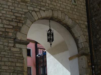 A close-up of a stone archway with a white plastered interior, featuring a hanging black metal lantern and a pink building visible through the opening.