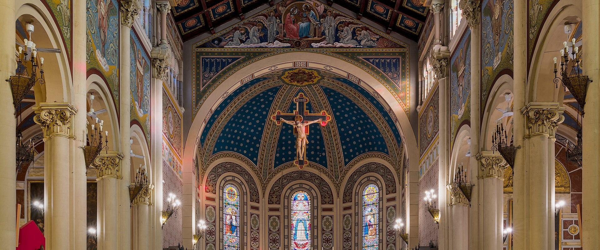 The interior of a grand cathedral featuring ornate columns, colorful frescoes, a large crucifix, and stained glass windows.