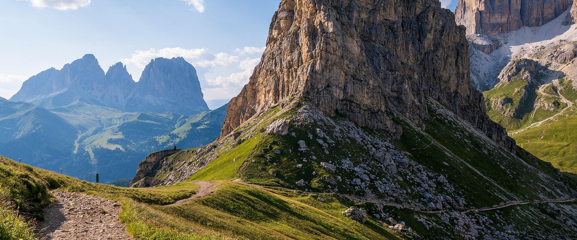 A dirt hiking trail winds through a high-altitude alpine meadow with dramatic limestone peaks, including the distinctive Sella Group and Marmolada visible in the background under a partly cloudy sky.