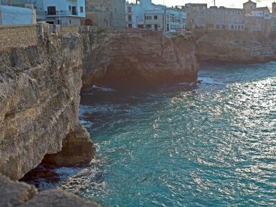 A view of steep limestone cliffs dropping into the turquoise Adriatic Sea, with traditional white and stone buildings perched along the cliff edge in the background.