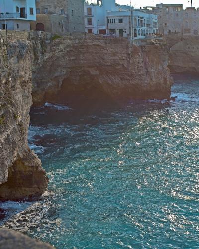A view of steep limestone cliffs dropping into the turquoise Adriatic Sea, with traditional white and stone buildings perched along the cliff edge in the background.