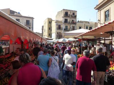 A crowded outdoor market scene with vendors selling produce under striped awnings, surrounded by multi-story buildings with balconies and shuttered windows, and people browsing or walking through the aisles.