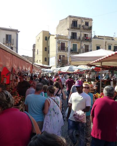 A crowded outdoor market scene with vendors selling produce under striped awnings, surrounded by multi-story buildings with balconies and shuttered windows, and people browsing or walking through the aisles.