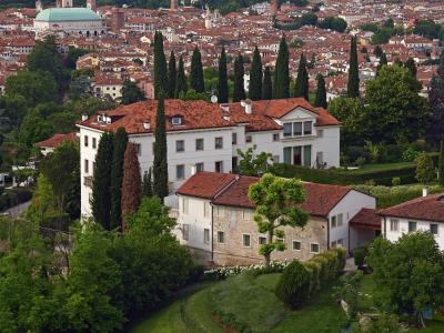 A high-angle view from a hillside showing white buildings with terracotta roofs and cypress trees in the foreground, overlooking the city of Vicenza with its distinctive green-domed Basilica of San Giorgio Maggiore and other historic architecture