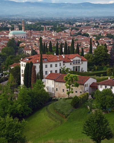 A high-angle view from a hillside showing white buildings with terracotta roofs and cypress trees in the foreground, overlooking the city of Vicenza with its distinctive green-domed Basilica of San Giorgio Maggiore and other historic architecture