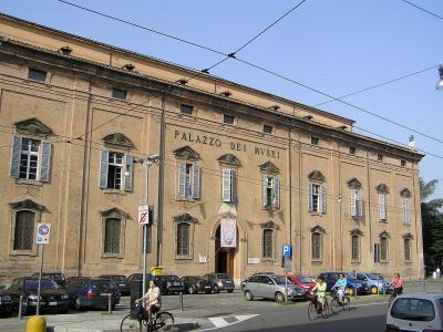 A large, historic brick building with the inscription "PALAZZO DEI MVSEI" on its facade, featuring shuttered windows, parked cars, and cyclists in the foreground.