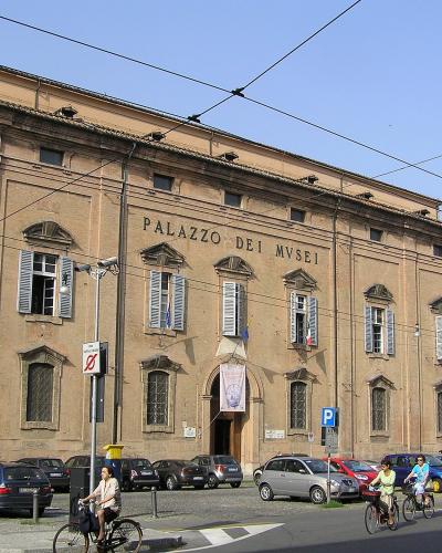 A large, historic brick building with the inscription "PALAZZO DEI MVSEI" on its facade, featuring shuttered windows, parked cars, and cyclists in the foreground.