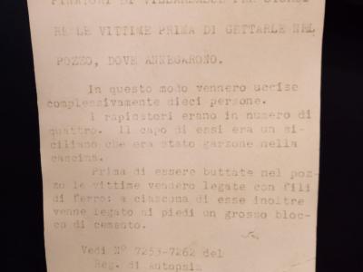 A close-up of a typed Italian museum label describing a wooden stave used by robbers in the Villarbasse massacre to stun victims before drowning them in a well, with autopsy registry numbers cited.