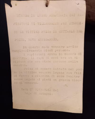 A close-up of a typed Italian museum label describing a wooden stave used by robbers in the Villarbasse massacre to stun victims before drowning them in a well, with autopsy registry numbers cited.