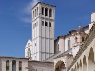 The image shows the white stone facade of the Basilica of San Francesco in Assisi, Italy, with its distinctive bell tower and rose window, alongside a modern bronze equestrian statue on a grassy lawn.
